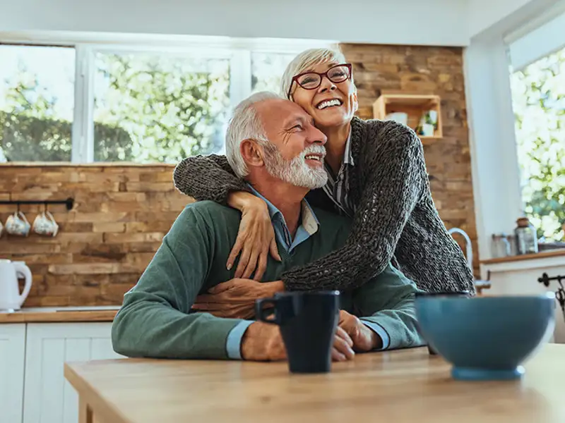 A mature couple embraces at the table in their kitchen.