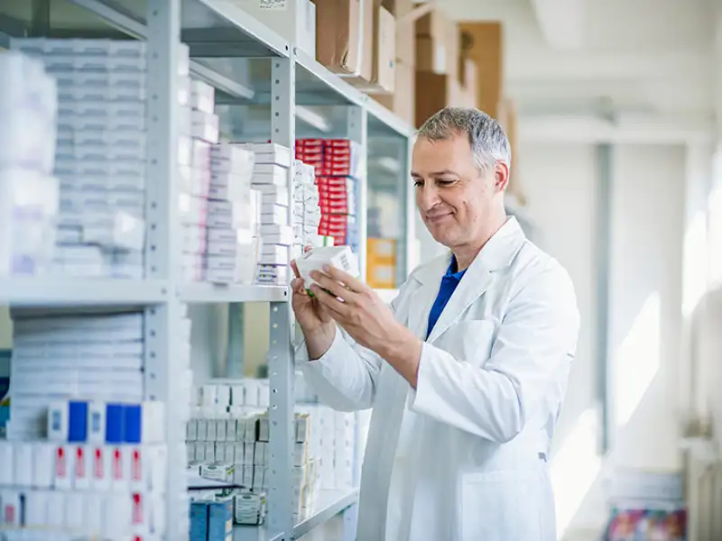 A pharmacist studies the packaging on a box in a large stockroom with many shelves of products.