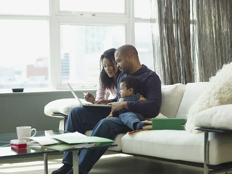 A young couple seated on a sofa with a toddler review papers and information on a laptop computer.