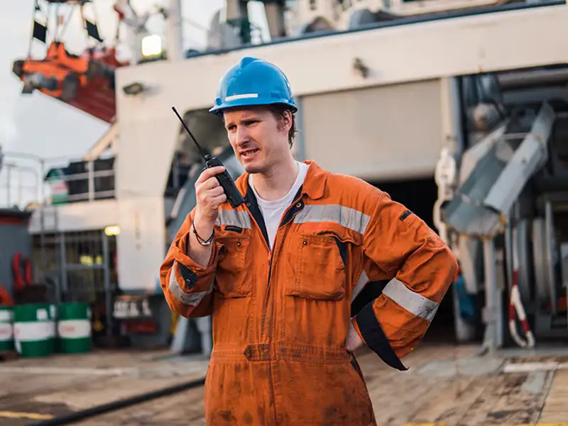 A marine worker in an orange jumpsuit and blue hardhat talks on a walkie-talkie.