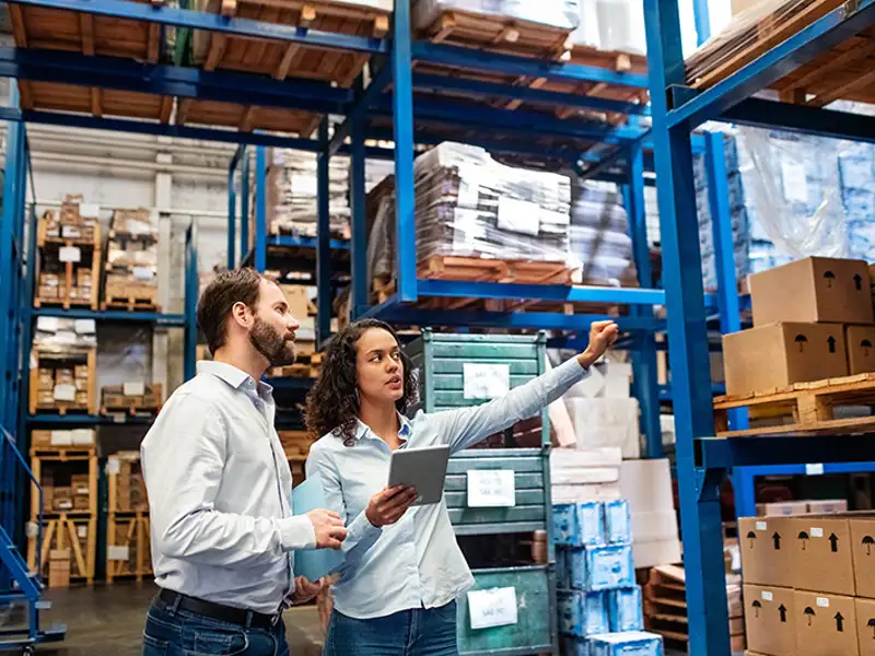 Two warehouse workers reference an electronic tablet as they inspect many shelves of boxes.
