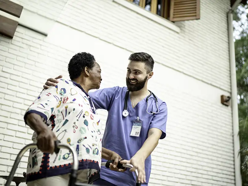 A medical professional assists a mature patient in moving while outdoors with a walker.