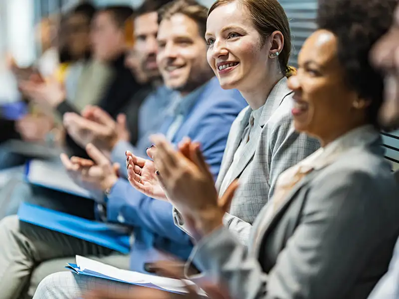 A row of seated audience members in business attire clap during a presentation.