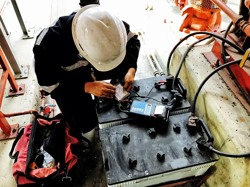 A technician in a black jumpsuit and white hardhat tests large batteries.