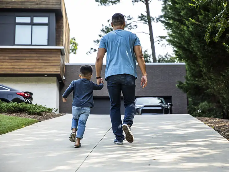 A father and young son hold hands as they walk up the driveway of their home.