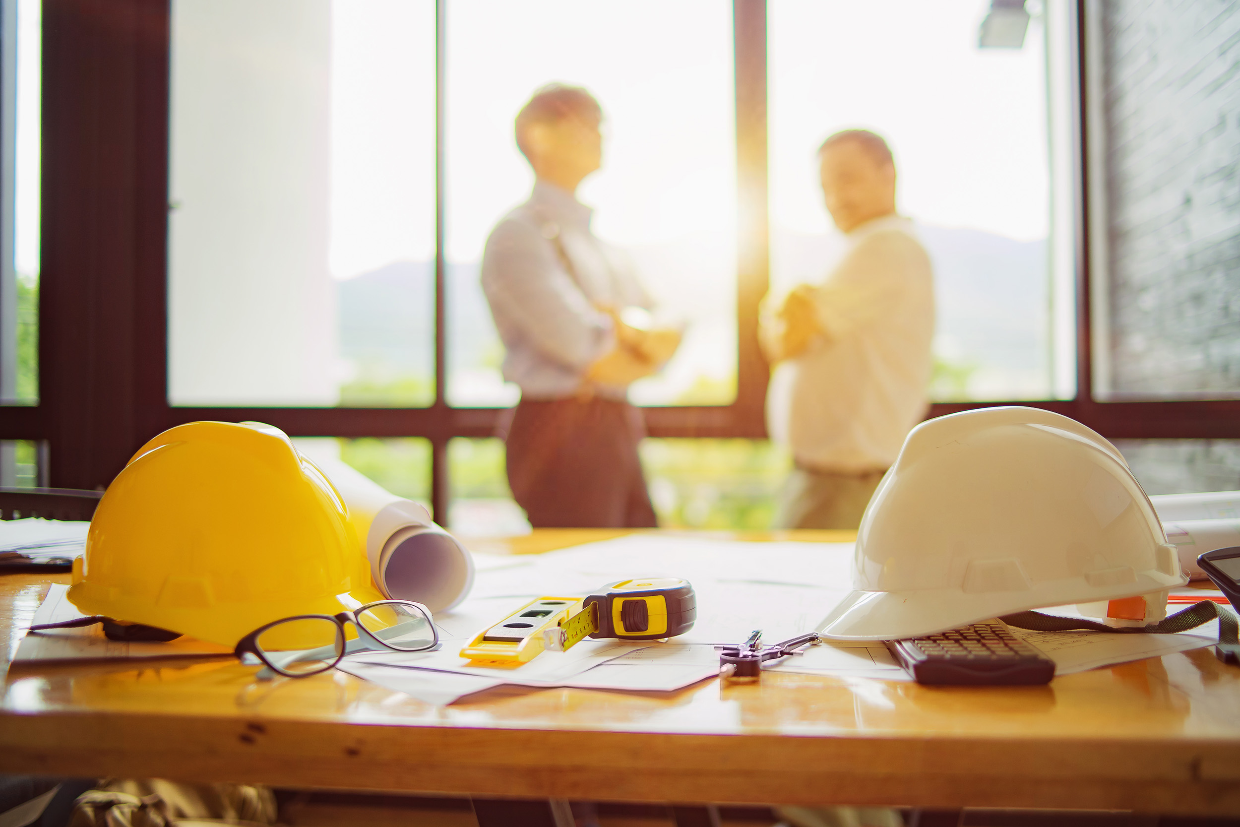 Two hardhats site on a table with construction administrators in the background.