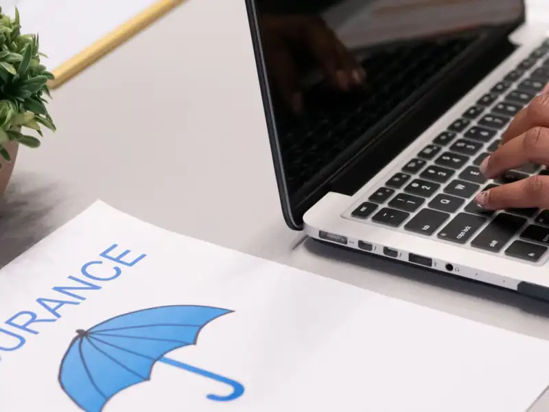 Closeup of a person on a computer with a piece of paper next to it with the word "insurance" and a picture of an umbrella.
