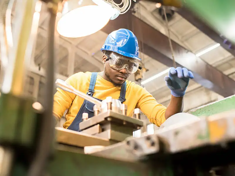 A worker in overalls and a blue hardhat works on a project in a manufacturing shop.