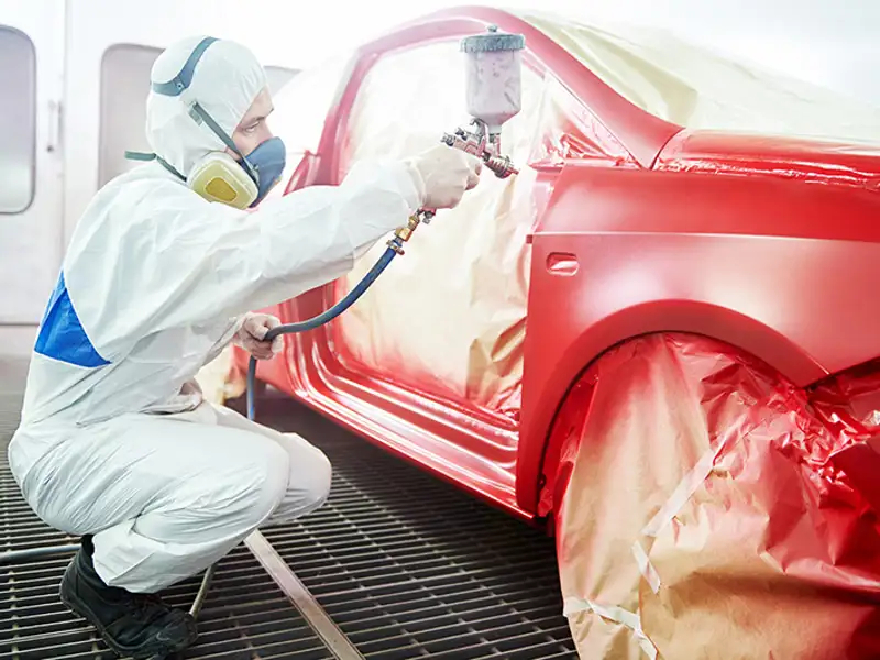 A technician in protective gear lays a coat of red paint on a vehicle with a spray gun.