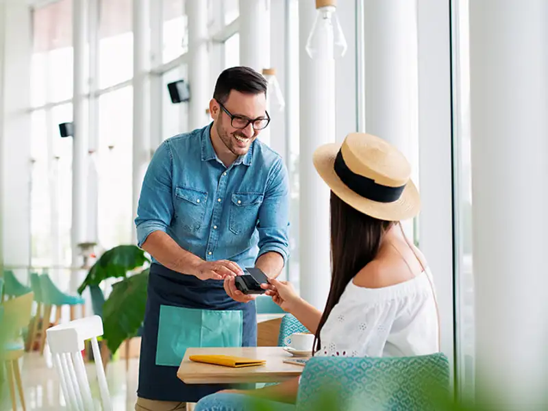 A smiling waiter processes the credit card payment of a young woman in a bright cafe.
