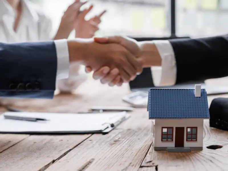 Two business people closing a deal shaking hands with a miniature house in the foreground.