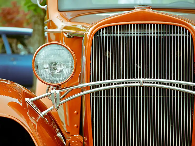 A view of the front grill and headlight of a pristine classic car painted burnt orange.