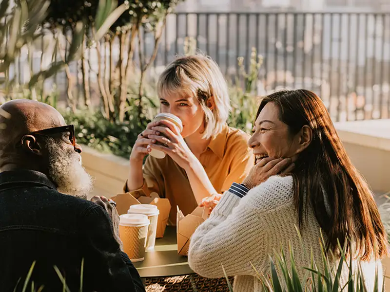 A trio of friends laugh and talk over coffee at an outdoor table in the morning.