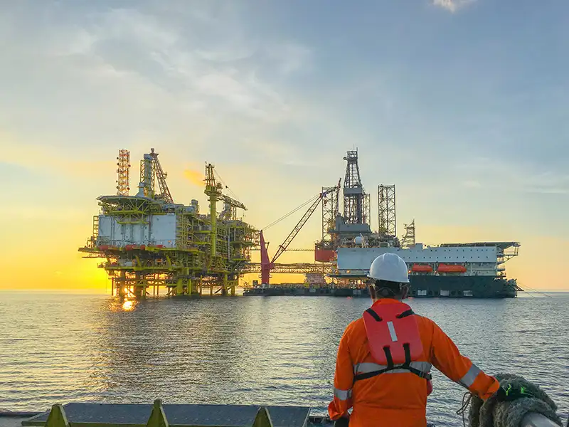 A worker in an orange jumpsuit and white hardhat looks out over the water at an offshore platform.
