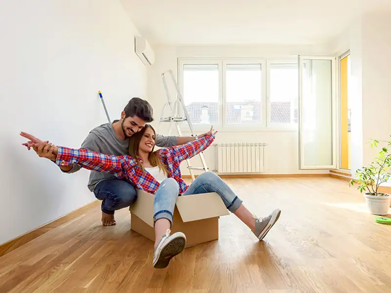 A young couple celebrates in an empty room of their new home by giddily sitting in a large moving box.