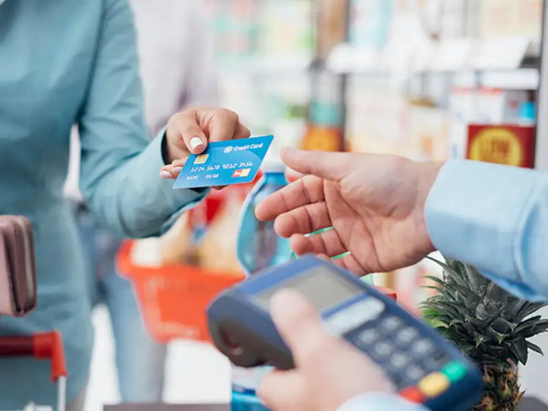 A store patron uses a credit card to purchase a pineapple at a grocery checkout.