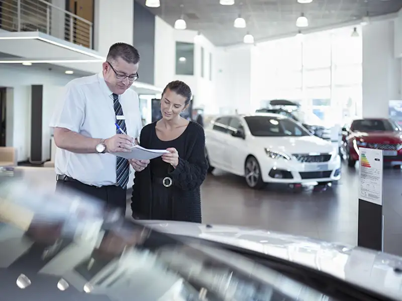 A woman reviews the paperwork of her new vehicle purchase with a sales agent on the showroom floor.