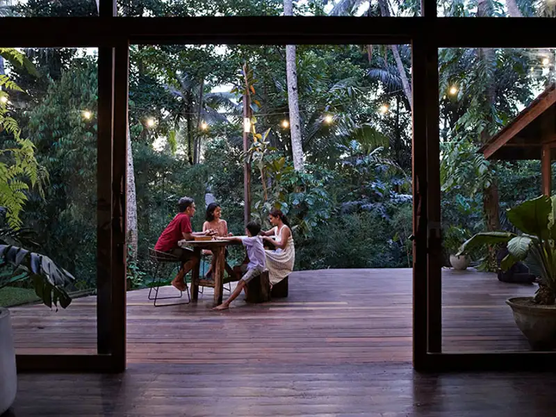 A casually-dressed family converses at a table on the wooden deck of a home in a lush forest.