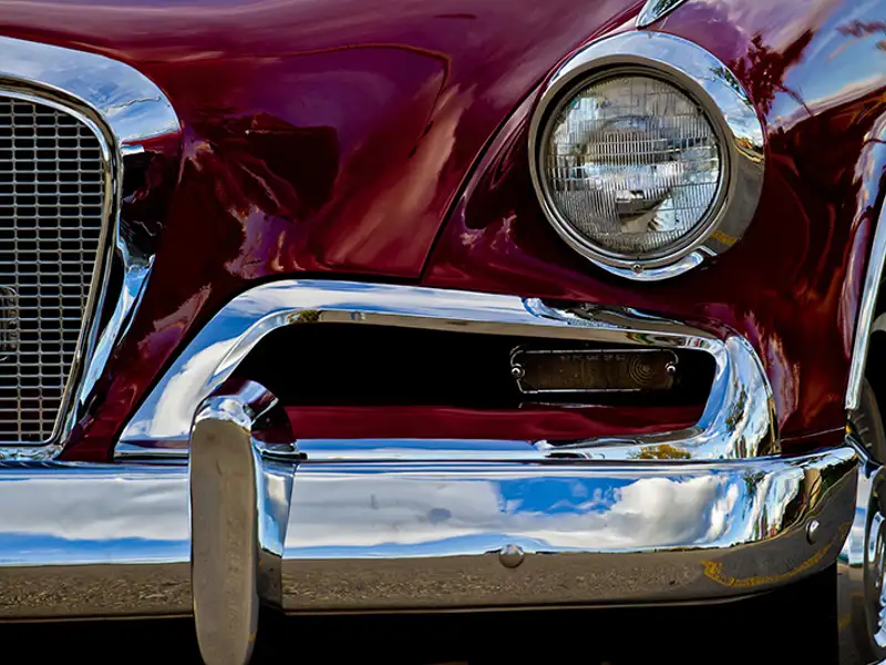 Closeup of the chrome bumper, grill and left headlight of a pristine burgundy-coloured classic car.