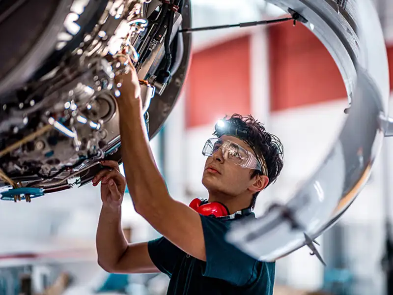 A young technician in safety glasses services a large aircraft engine.