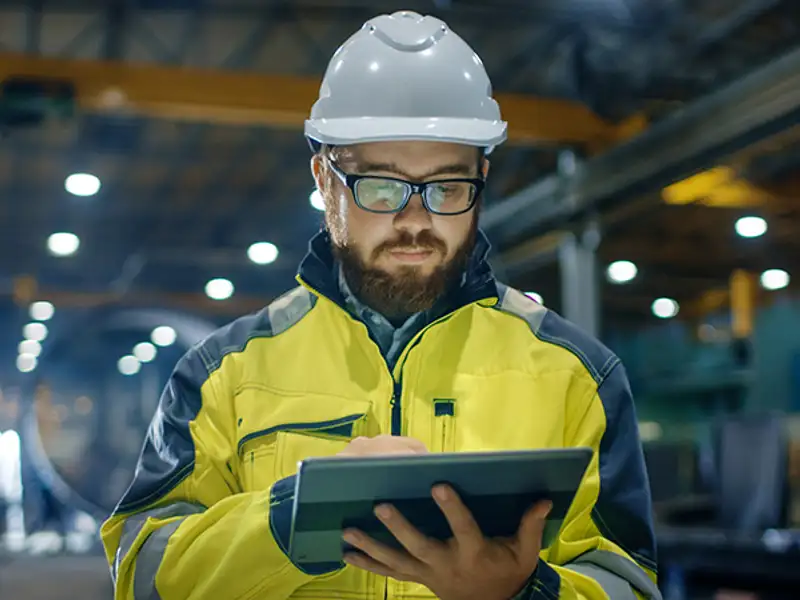 A technician in a hard hat studies information on a tablet in a large facility.