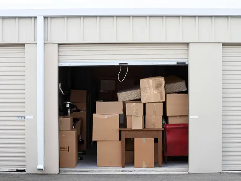 An open rolling door reveals stacked cardboard boxes in a unit at a storage facility.