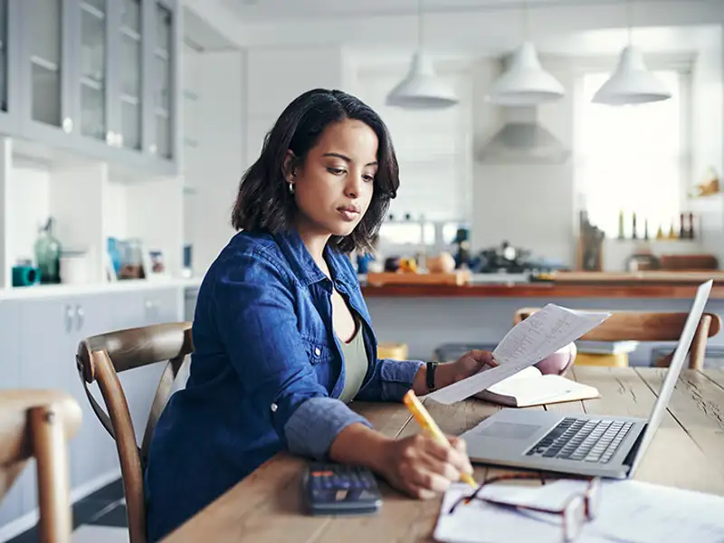 A woman in a blue denim shirt works reviews papers in front of a laptop computer at a kitchen table.